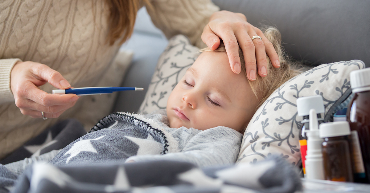 Sick child, toddler boy lying on the couch in living room with a fever, mom cheching his temperature, resting at home; blog: cold versus flu how to tell the difference