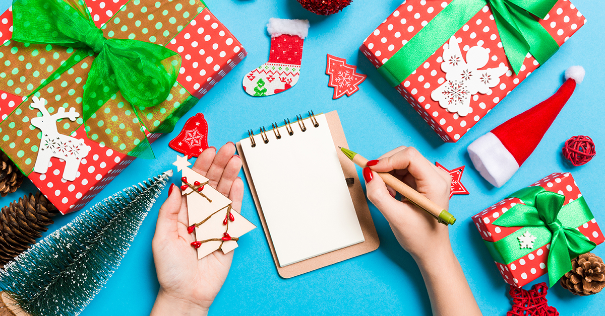 Top view of female hand making some notes in noteebok on blue background. New Year decorations and toys. Christmas time concept; blog: 14 Non-Toy Gift Ideas for Kids