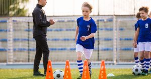 Young Happy Blonde Soccer Player Running with Ball on Training. Coach of Youth Football Team Explaining Drill Exercise in the Background. Caucasian Soccer Players on Practice Session; blog: Sports Physicals for Kids: What to Expect