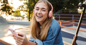 Image of a laughing cheery smiling young teenage girl outside in park listening music with headphones holding mobile phone showing tongue; blog: stages of adolescence