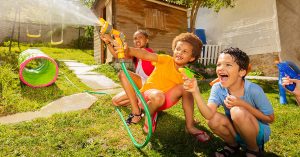 Group of happy kids scram smile squat and shoot with water guns, garden hose playing game on backyard; blog: 8 Healthy Activities for Kids to Do While School’s Out