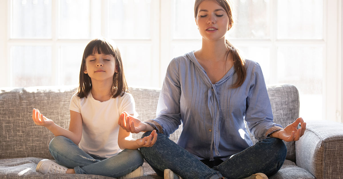 Calm smiling mother with cute little daughter doing yoga exercise at home, pretty girl and attractive mum with closed eyes sitting in lotus pose on couch, relaxing together, family exercise; blog: 9 Mental Health Tips for Kids in Stressful Times