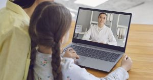 Family doctor online. Mother and girl kid talking consults a doctor using a laptop while sitting at home on the couch. Online medical consultation; blog: 7 Ways to Prep for Your Child’s Telemedicine Visit