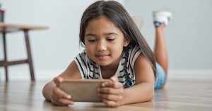 Little asian girl lying on floor using mobile phone to play. Multiethnic female child watching cartoon on smartphone at home. Childhood and moder technology concept; blog: 8 Tips for Limiting Screen Time for Kids