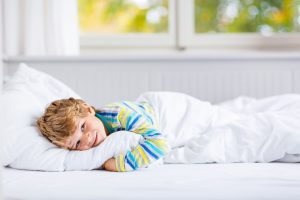 Adorable happy little kid boy after sleeping in his white bed in colorful nightwear near big window with green and yellow autumn foliage. Funny happy child playing and smiling. Family, vacation, childhood concept