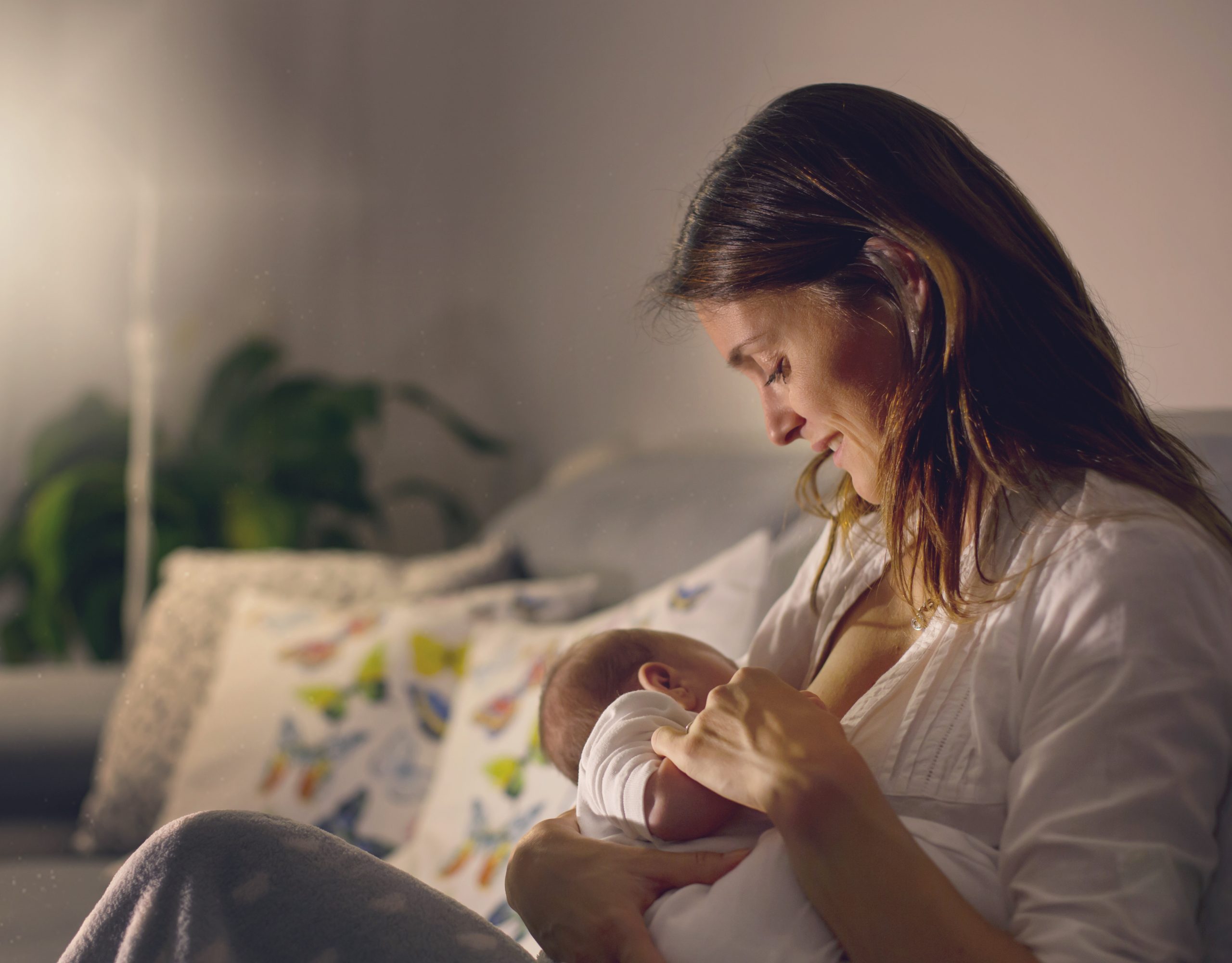 Young beautiful mother, breastfeeding her newborn baby boy at night, dim light. Mom breastfeeding infant